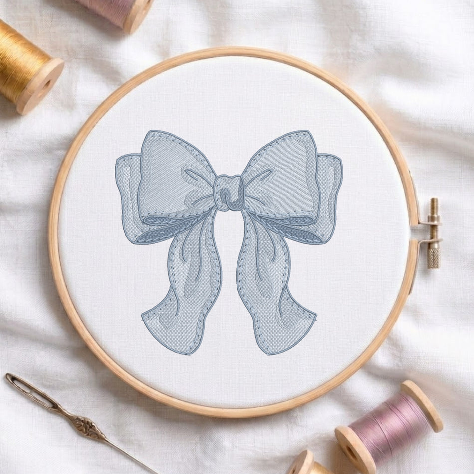 A wooden embroidery hoop holding white fabric with a stitched bow design in pale blue-gray. Surrounding the hoop are spools of thread in gold, pink, and lavender, plus a metal needle on a soft white fabric background.