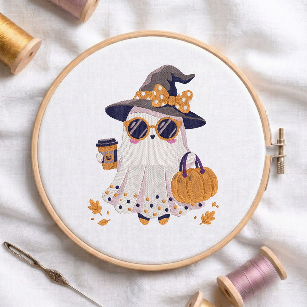 A circular embroidery hoop holds a white fabric with a stylized embroidered ghost wearing a witch hat, sunglasses, holding a pumpkin, and a coffee cup. The hoop is styled on a white fabric background with spools of thread, a needle, and autumn leaves.