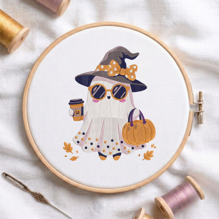 A circular embroidery hoop holds a white fabric with a stylized embroidered ghost wearing a witch hat, sunglasses, holding a pumpkin, and a coffee cup. The hoop is styled on a white fabric background with spools of thread, a needle, and autumn leaves.