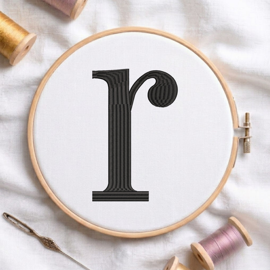 A wooden embroidery hoop holding white fabric with a large black embroidered letter 'r' in a stylized serif font. The hoop is photographed on white fabric with spools of thread and a needle around it.