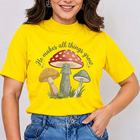 A woman wearing a bright yellow graphic t-shirt with a mushroom design and the text 'He makes all things grow.' paired with blue jeans, photographed against a light gray background.