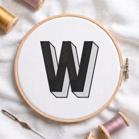 A wooden embroidery hoop holding white fabric with a large black letter W stitched in the center. A metal needle lies at the lower left, and spools of thread in gold, pink, and white are arranged around the hoop on a white fabric background.