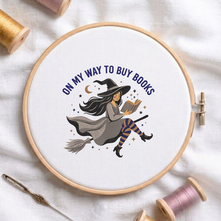 On my way to buy books witch reading on a broom machine embroidery design displayed in a wooden hoop on white fabric with thread spools and a needle   