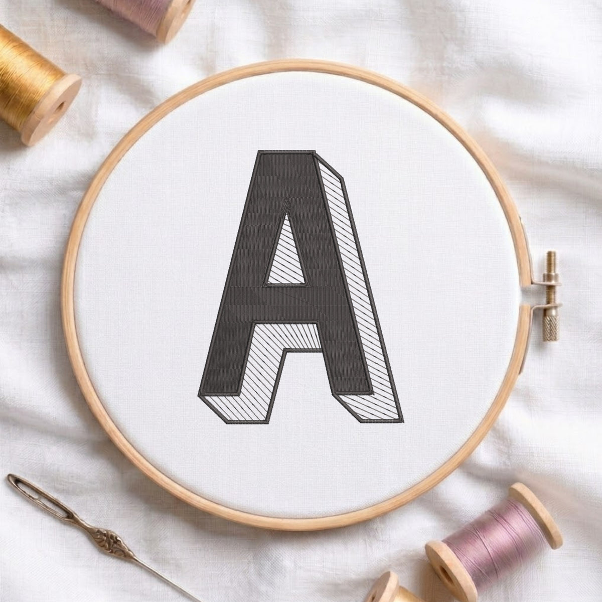 A wooden embroidery hoop holding white fabric with a large outlined letter A, surrounded by spools of thread and a needle on a white fabric background.