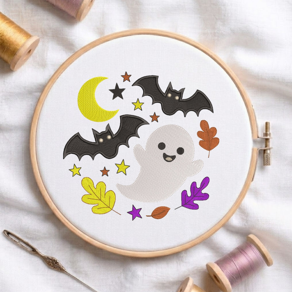 A circular embroidery hoop on white fabric, featuring a cute Halloween design with a white ghost, two black bats, a yellow crescent moon, yellow and orange stars, and autumn leaves. Spools of thread and a needle are arranged around the hoop.