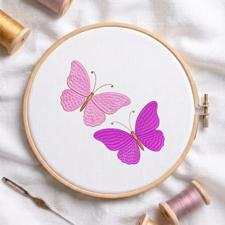 A circular embroidery hoop holding white fabric with two embroidered butterflies, one pink and one purple, surrounded by spools of thread and a needle on a white fabric background.