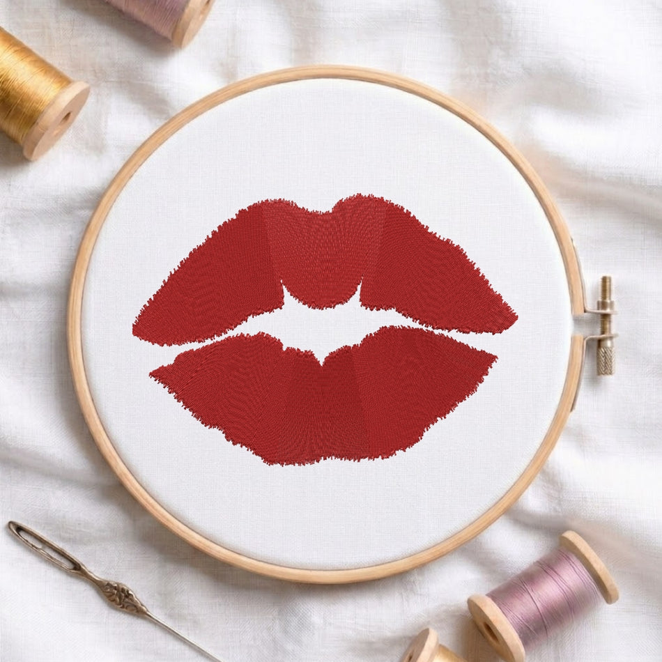 The red lips embroidery design secured in a wooden embroidery hoop, displayed with embroidery thread spools and a needle on a white fabric background.