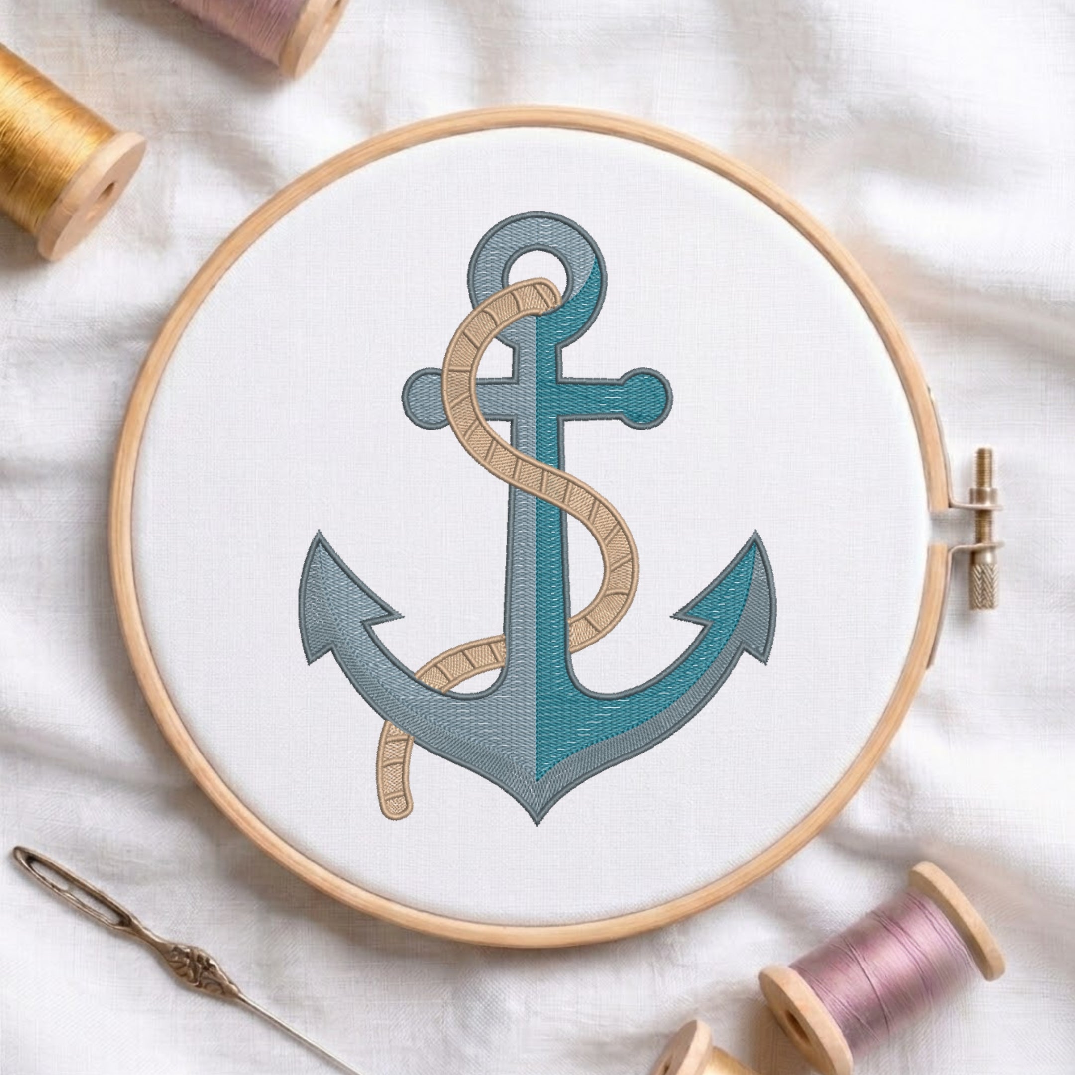 A wooden embroidery hoop holds white fabric with a stitched anchor design in teal and gray, wrapped with a beige rope motif. A needle and thread spools are arranged around it on a white fabric background.