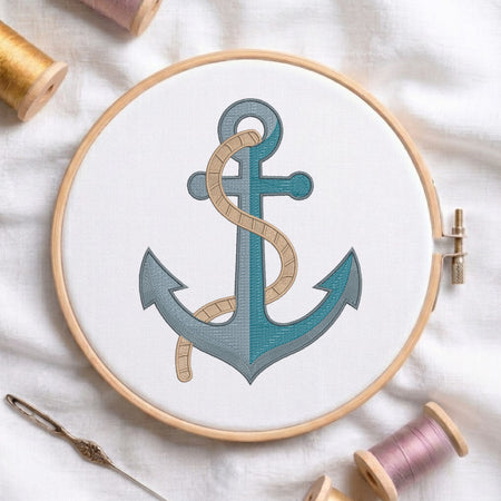 A wooden embroidery hoop holds white fabric with a stitched anchor design in teal and gray, wrapped with a beige rope motif. A needle and thread spools are arranged around it on a white fabric background.