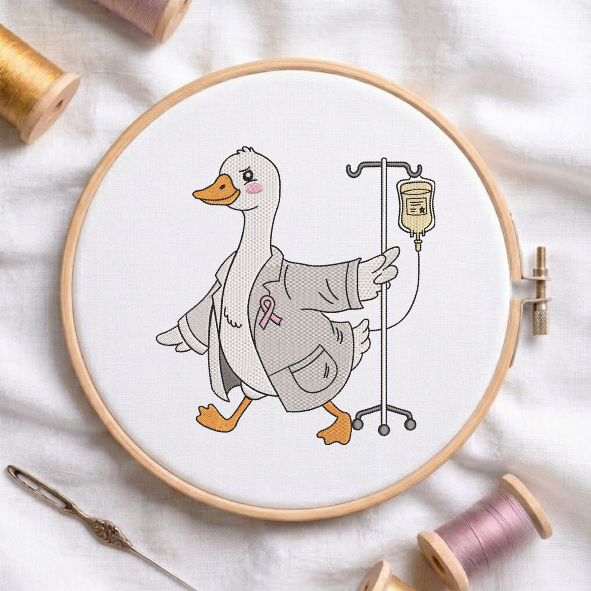An embroidered design of a cute white duck wearing a doctor's lab coat and a pink awareness ribbon, walking with an IV pole, stitched inside a wooden hoop on white fabric with sewing threads.