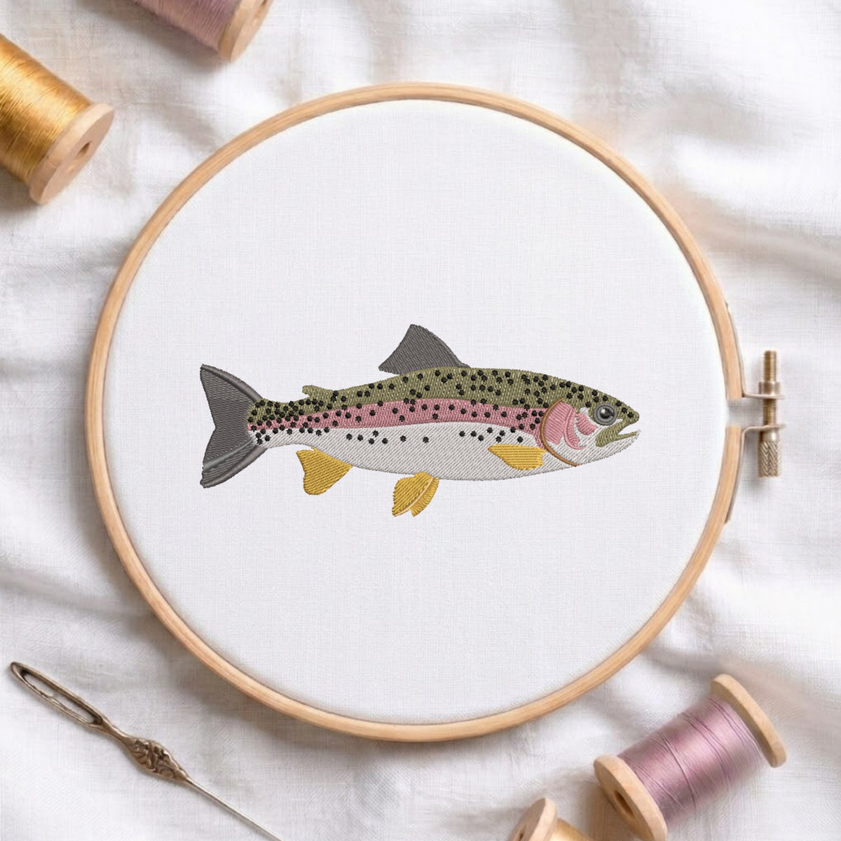 A round embroidery hoop holding white fabric with a stitched rainbow trout design in muted green, pink, yellow, and gray tones. The hoop is photographed on white fabric with spools of thread and a needle around it.