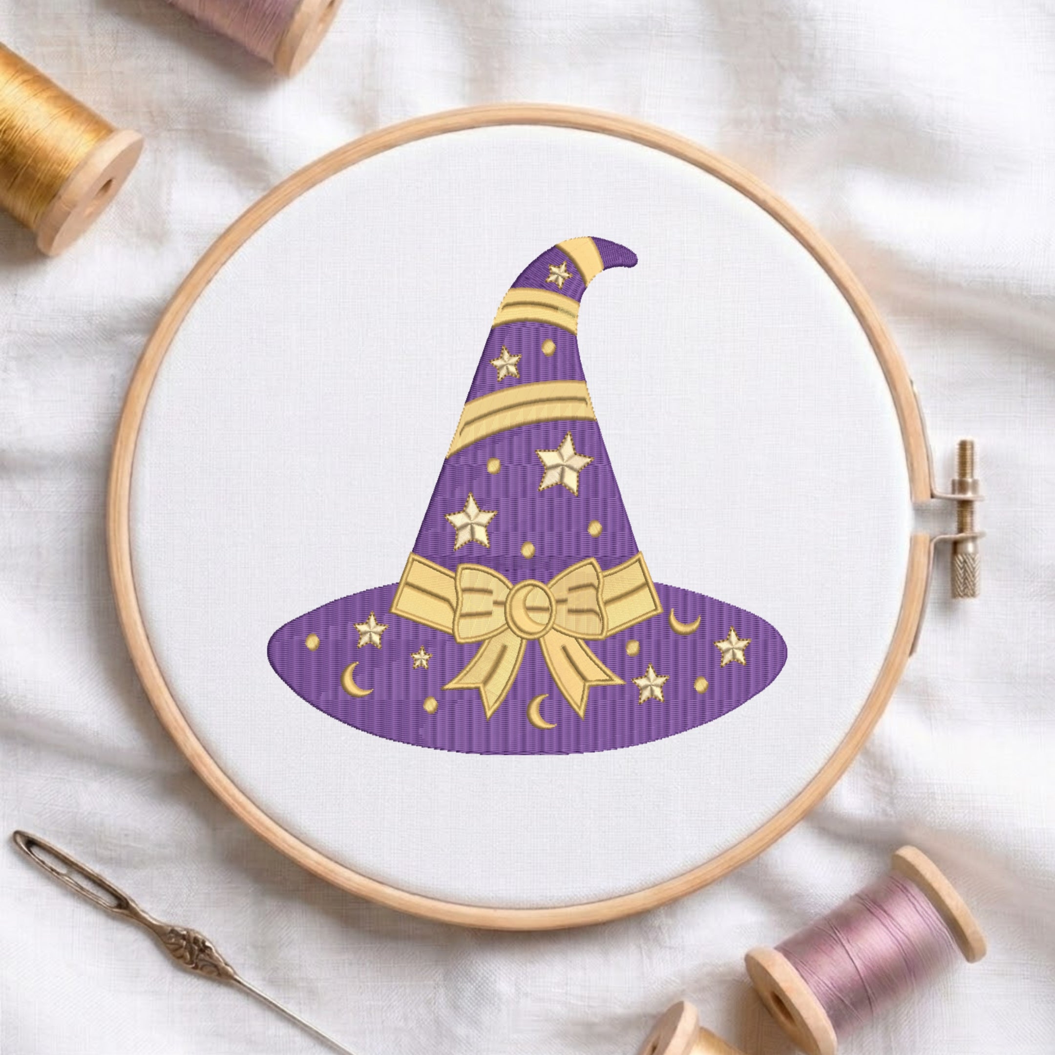 A circular embroidery hoop holding white fabric with a purple witch hat design outlined in gold, surrounded by spools of thread and a needle on a white cloth background.