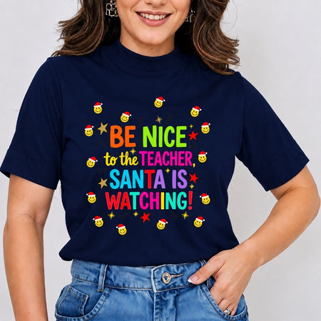 A woman wearing a navy blue graphic t-shirt with the text 'BE NICE to the TEACHER, SANTA IS WATCHING!' in colorful letters, surrounded by small yellow smiley faces wearing Santa hats and stars. She is wearing blue jeans against a plain light gray background.