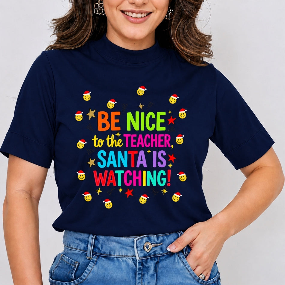 A woman wearing a navy blue graphic t-shirt with the text 'BE NICE to the TEACHER, SANTA IS WATCHING!' in colorful letters, surrounded by small yellow smiley faces wearing Santa hats and stars. She is wearing blue jeans against a plain light gray background.