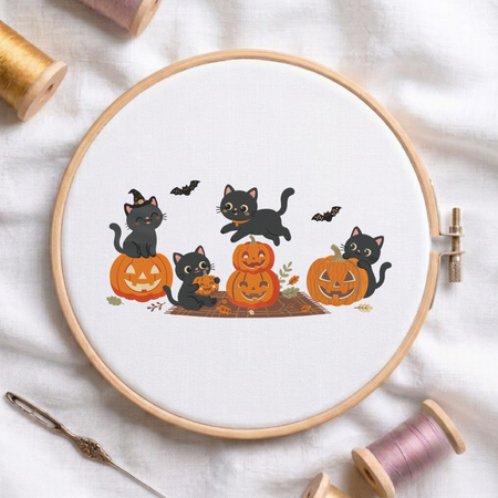 Cute black cats playing with jack-o-lantern pumpkins and bats Halloween machine embroidery design shown in a wooden embroidery hoop on white fabric with thread spools and a needle