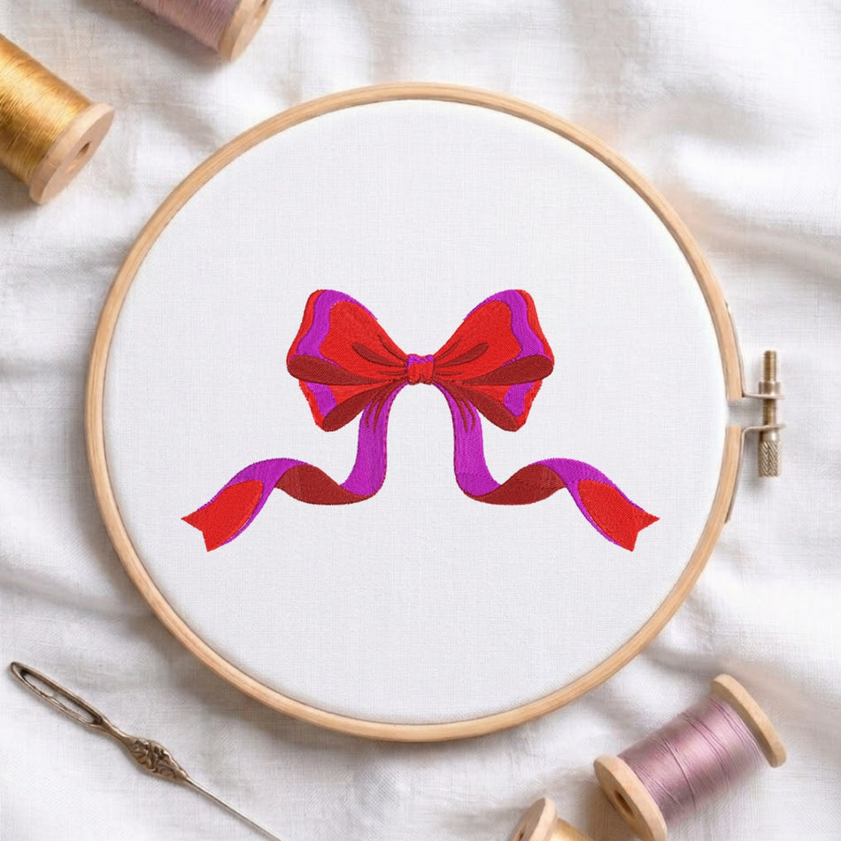 A circular embroidery hoop on white fabric, featuring a stylized red and purple bow design. Spools of thread and a needle are arranged around it on a white fabric background.