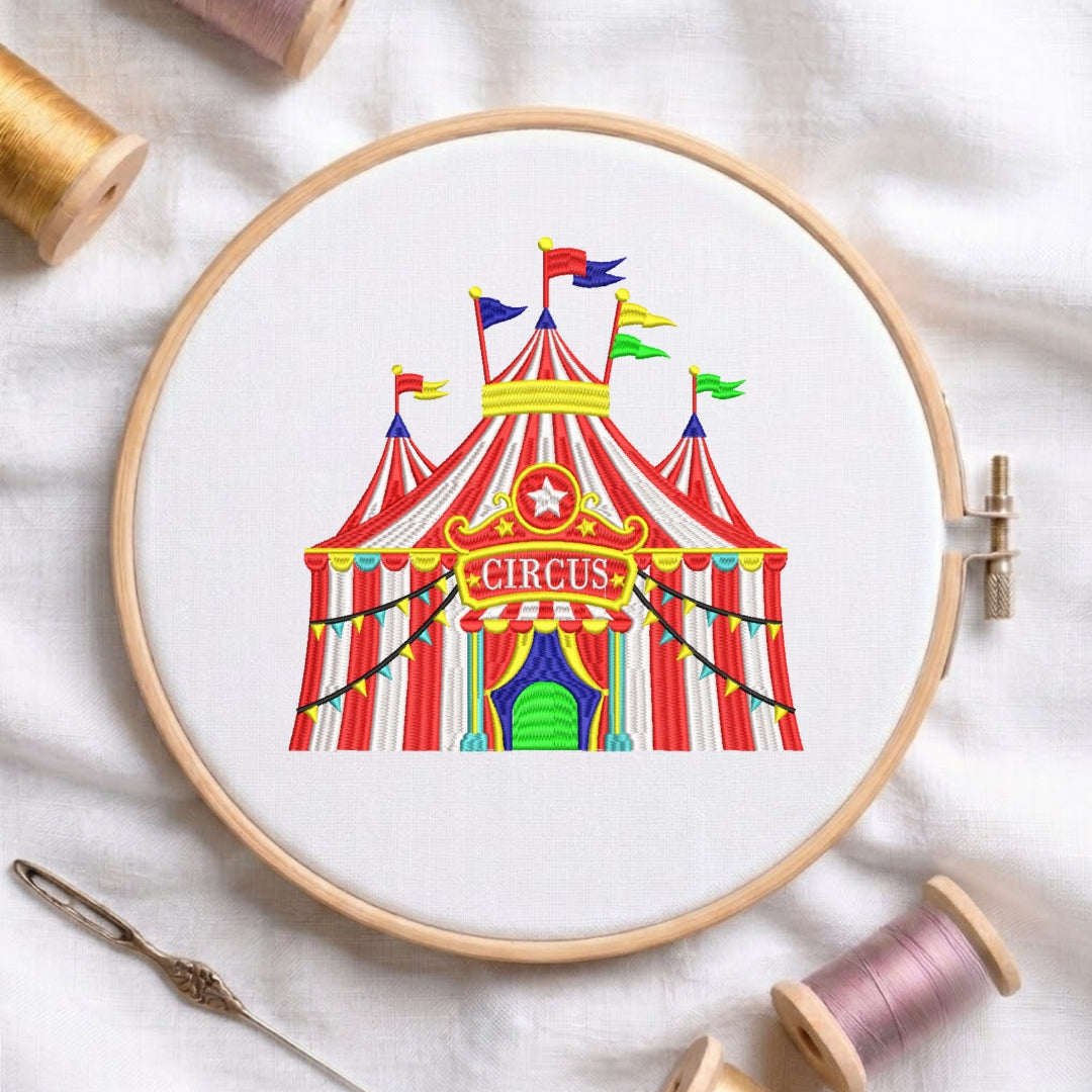 A circular embroidery hoop holding white fabric with a colorful circus tent design stitched inside. The hoop is styled on a white fabric background with spools of thread and a needle around it.