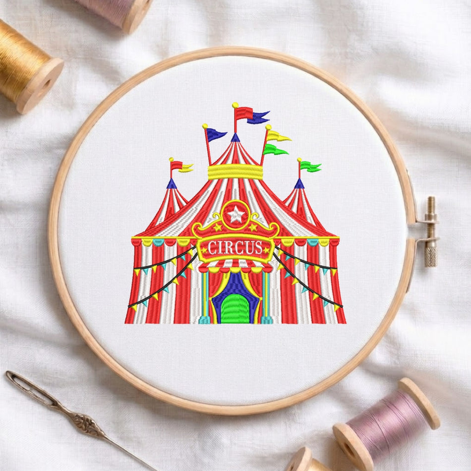 A circular embroidery hoop holding white fabric with a colorful circus tent design stitched inside. The hoop is styled on a white fabric background with spools of thread and a needle around it.