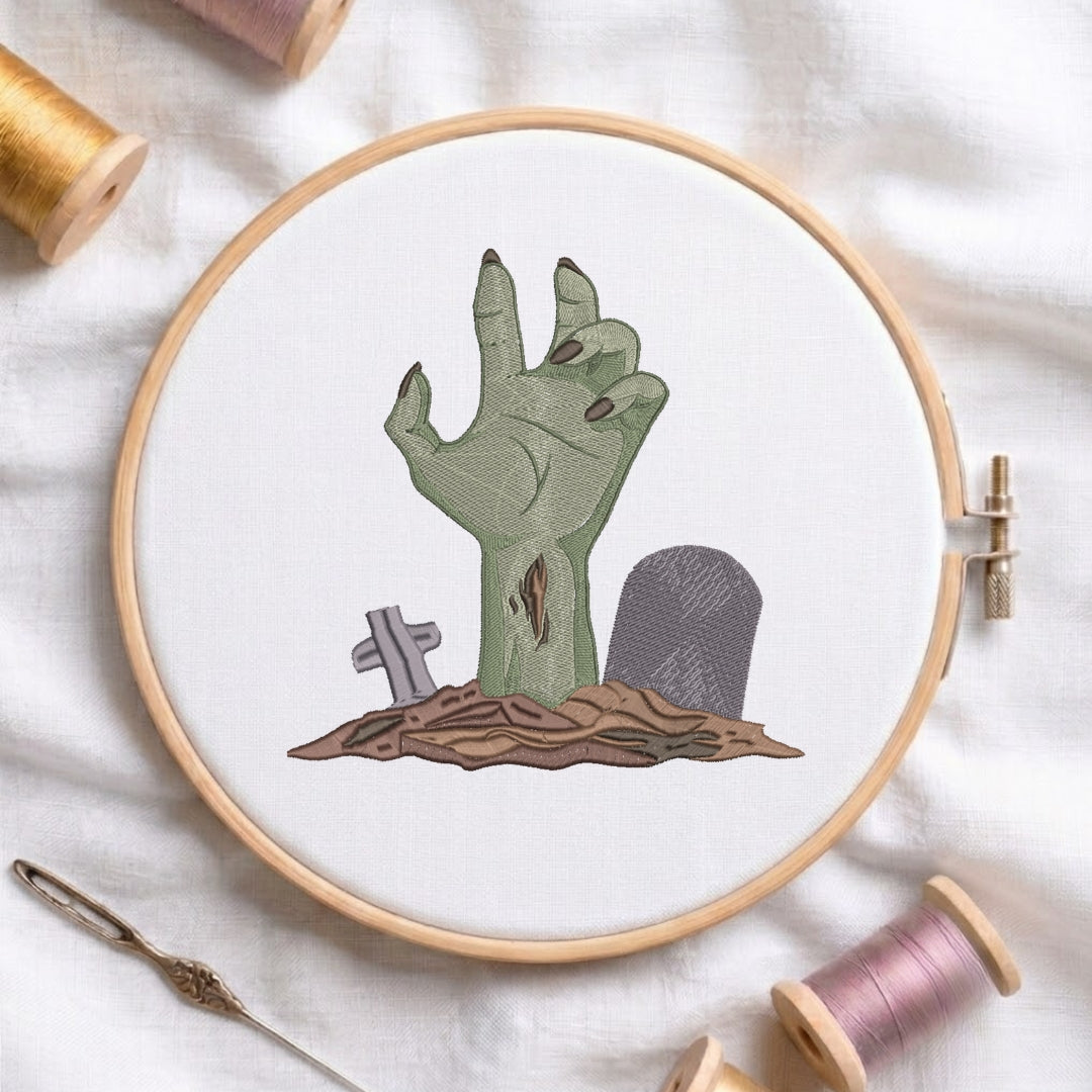 A circular embroidery hoop holding white fabric with an embroidered zombie hand rising from a patch of brown ground beside small gray tombstones. The hoop is styled on a white fabric background with spools of thread and a needle nearby.