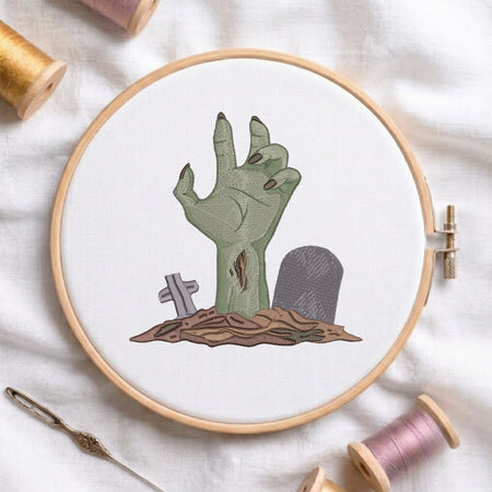 A circular embroidery hoop holding white fabric with an embroidered zombie hand rising from a patch of brown ground beside small gray tombstones. The hoop is styled on a white fabric background with spools of thread and a needle nearby.