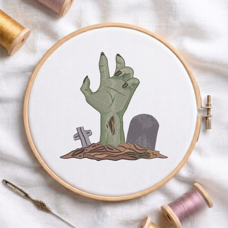 A circular embroidery hoop holding white fabric with an embroidered zombie hand rising from a patch of brown ground beside small gray tombstones. The hoop is styled on a white fabric background with spools of thread and a needle nearby.