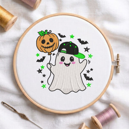 A white fabric stretched in a wooden embroidery hoop, featuring an embroidered cute ghost wearing a black and green baseball cap, holding a jack-o'-lantern pumpkin balloon, surrounded by bats and green and black stars.