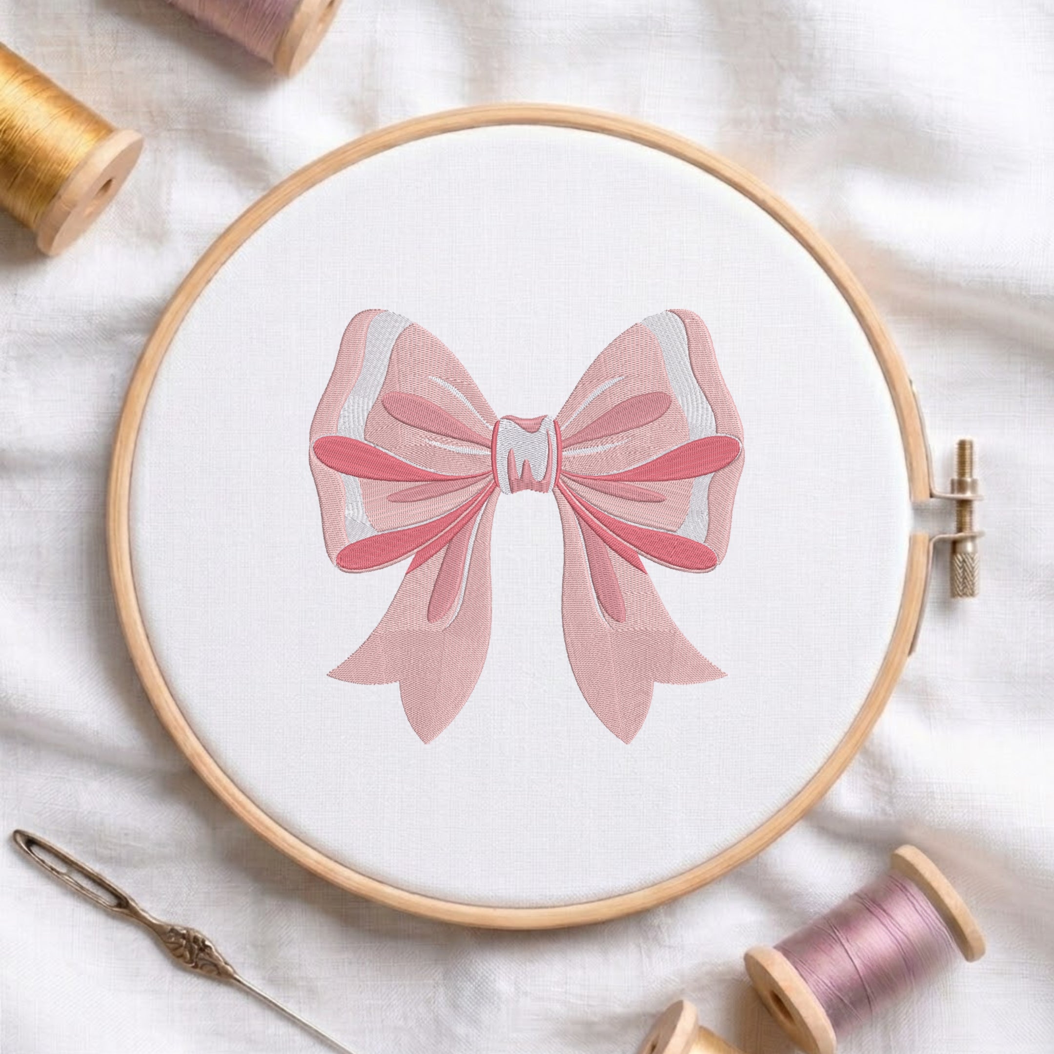 A wooden embroidery hoop holding white fabric with a pink bow design stitched in the center, surrounded by spools of thread and a needle on a white fabric background.