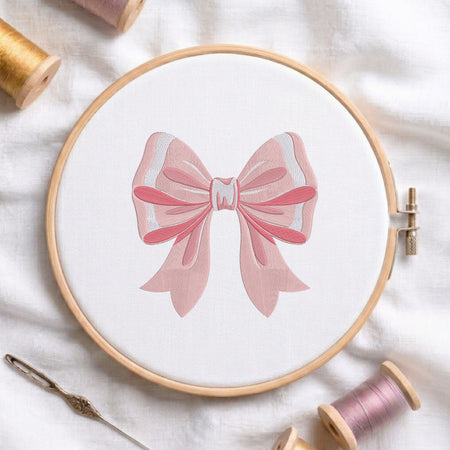 A wooden embroidery hoop holding white fabric with a pink bow design stitched in the center, surrounded by spools of thread and a needle on a white fabric background.