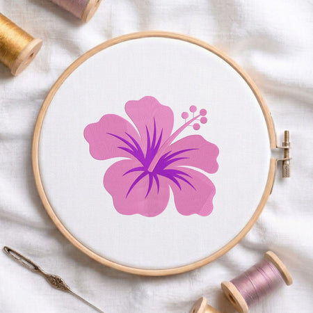 A wooden embroidery hoop holding white fabric with a large pink hibiscus flower design in the center. A metal needle lies at the lower left, and spools of thread are arranged around the hoop on a white fabric background.