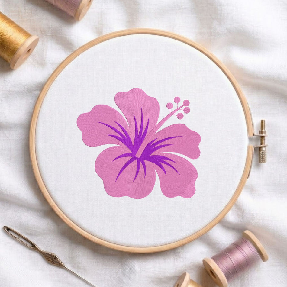 A wooden embroidery hoop holding white fabric with a large pink hibiscus flower design in the center. A metal needle lies at the lower left, and spools of thread are arranged around the hoop on a white fabric background.