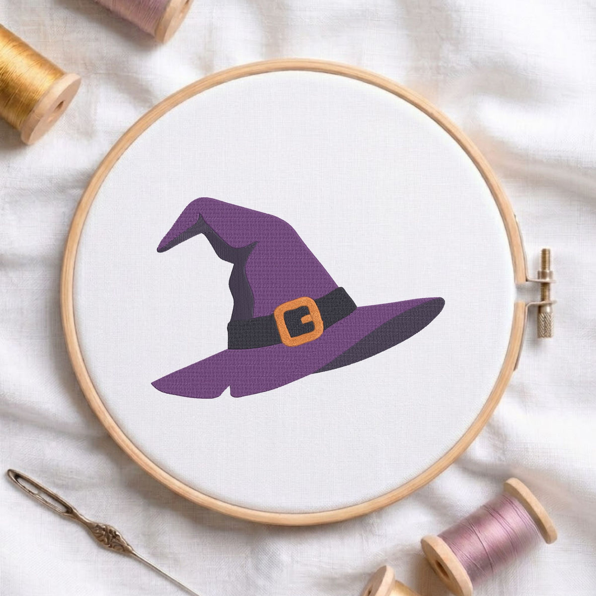 A circular embroidery hoop holding white fabric with a purple witch hat design, surrounded by spools of thread and a needle on a white fabric background.