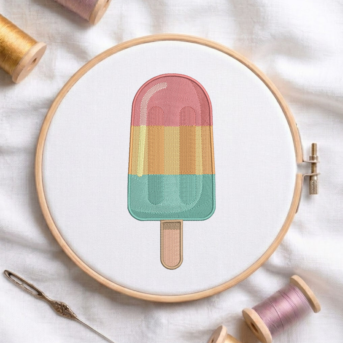 A circular embroidery hoop holds a colorful popsicle design stitched with pastel pink, yellow, and mint green threads on white fabric, surrounded by spools of thread and a needle.
