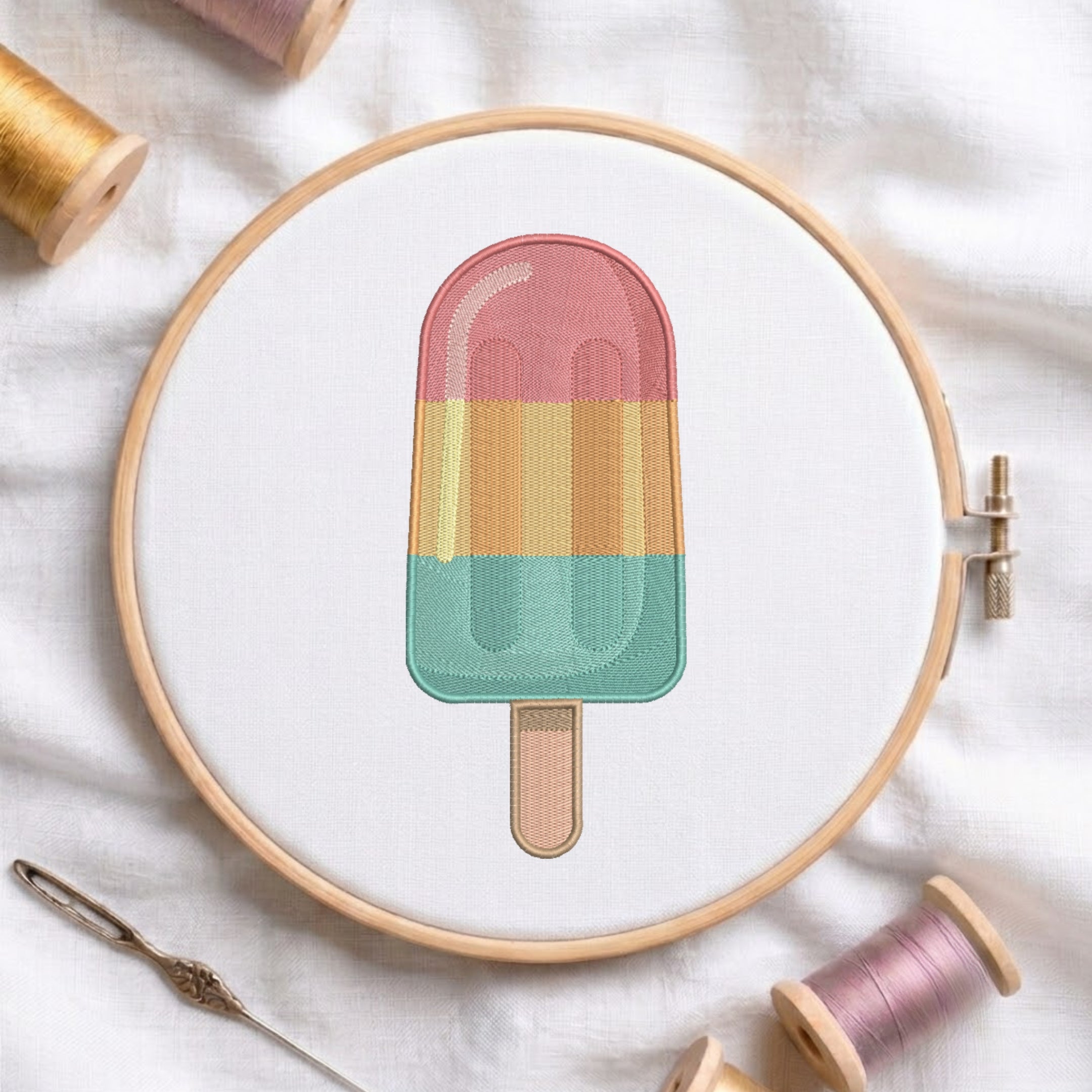 A circular embroidery hoop holds a colorful popsicle design stitched with pastel pink, yellow, and mint green threads on white fabric, surrounded by spools of thread and a needle.