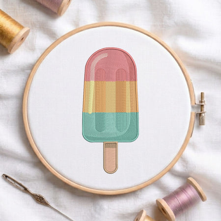 A circular embroidery hoop holds a colorful popsicle design stitched with pastel pink, yellow, and mint green threads on white fabric, surrounded by spools of thread and a needle.