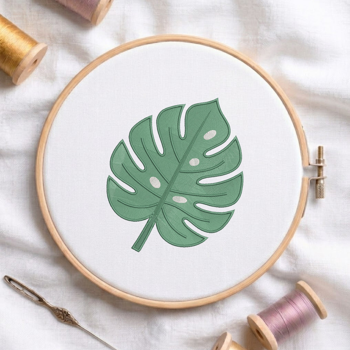 A wooden embroidery hoop holding white fabric with a green monstera leaf design, styled on a white cloth with thread spools and a needle around it.