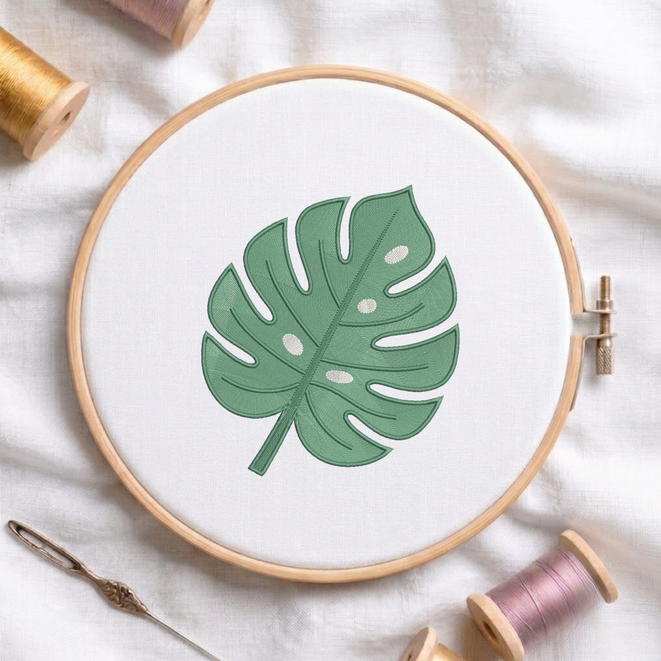 A wooden embroidery hoop holding white fabric with a green monstera leaf design, styled on a white cloth with thread spools and a needle around it.
