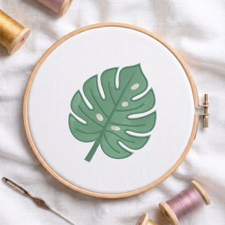 A wooden embroidery hoop holding white fabric with a green monstera leaf design, styled on a white cloth with thread spools and a needle around it.