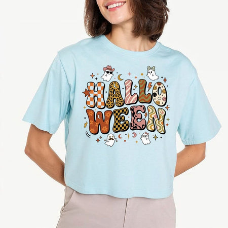 A woman wearing a light blue cropped t-shirt with a colorful 'HALLOWEEN' graphic featuring cartoon ghosts and patterned letters, paired with beige pants, against a plain white background.