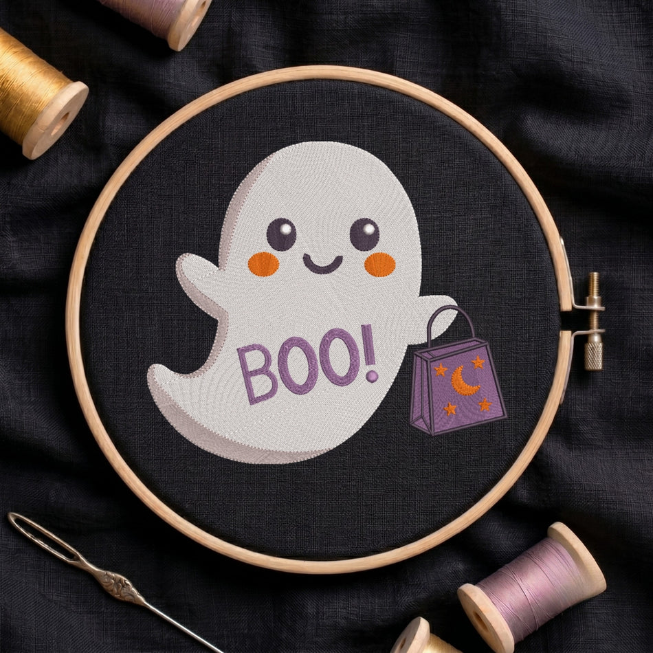 A circular embroidery hoop holding a black fabric with a cute white ghost design that says 'BOO!' in purple and holds a purple candy bag. Spools of thread and a needle are arranged around it on a dark fabric background.