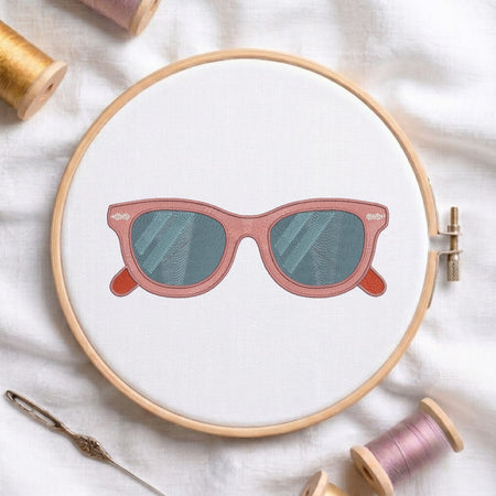 A circular embroidery hoop on white fabric, featuring a stitched illustration of pink sunglasses with blue-green lenses. Spools of thread and a needle are arranged around the hoop.