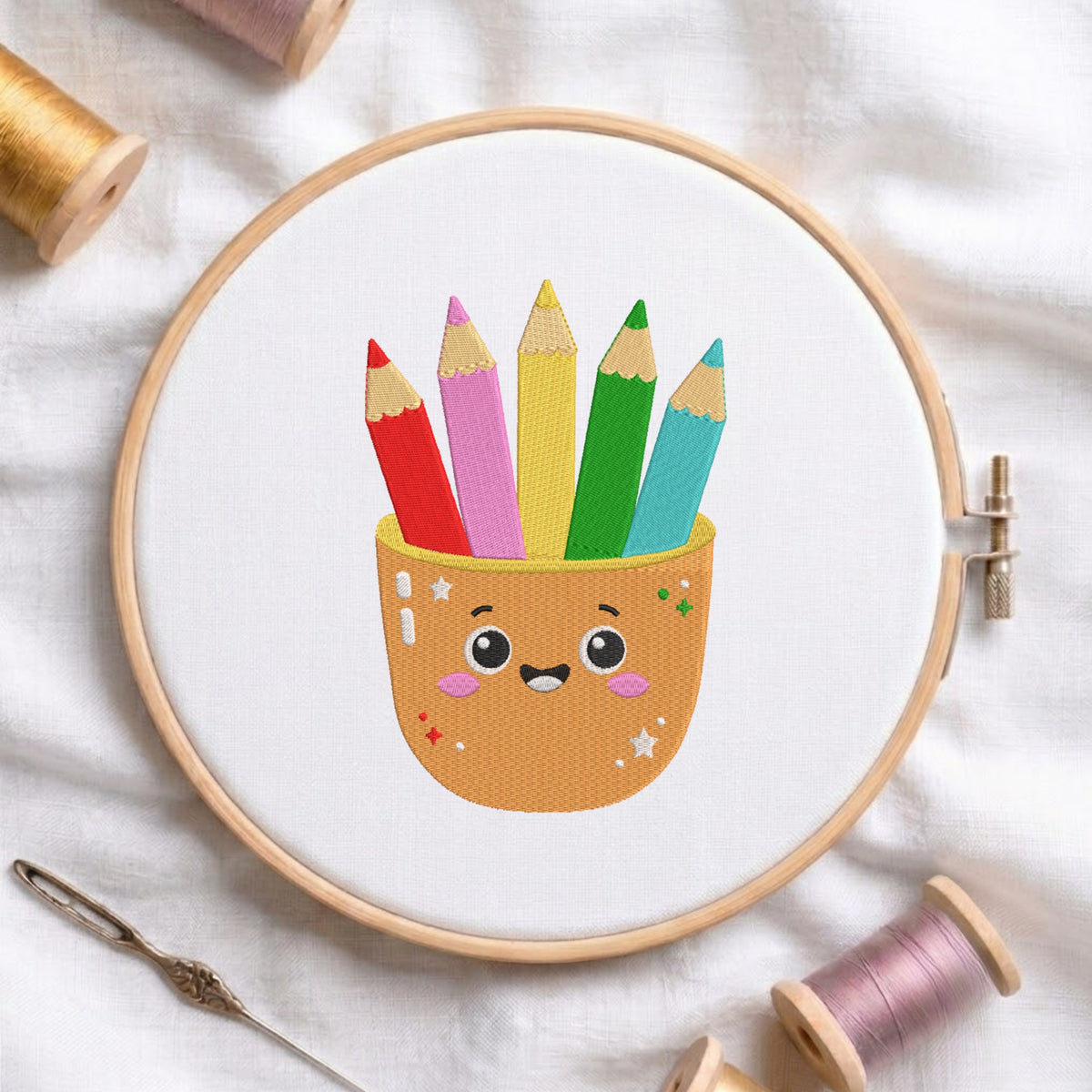 An embroidered design of a cute smiling kawaii cup holding colorful pencils in red, pink, yellow, green, and blue, stitched inside a wooden hoop on white fabric with sewing threads.