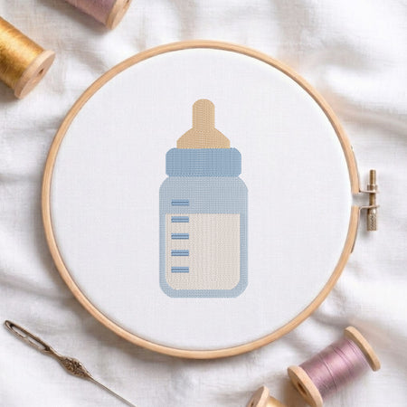 A baby bottle cross-stitch design held in a wooden embroidery hoop, surrounded by spools of thread and a needle on a white fabric background.