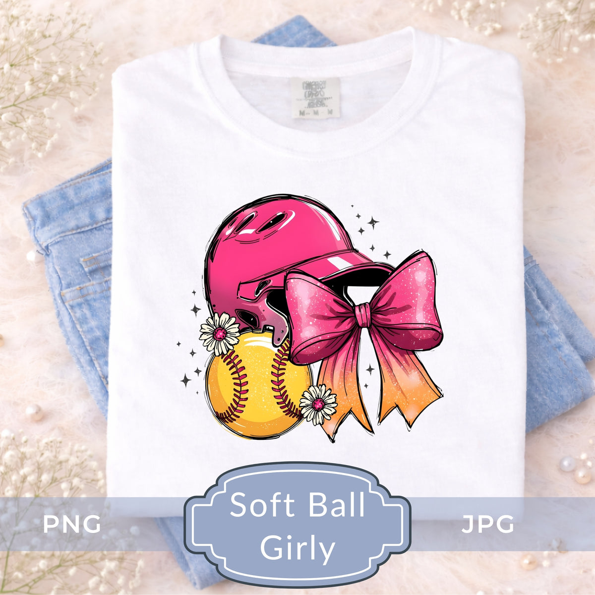 A folded white T-shirt displayed on top of light blue jeans, featuring a colorful graphic of a pink softball helmet, yellow softball, and pink bow with daisies. Text overlay at the bottom reads 'Soft Ball Girly' with PNG and JPG labels.