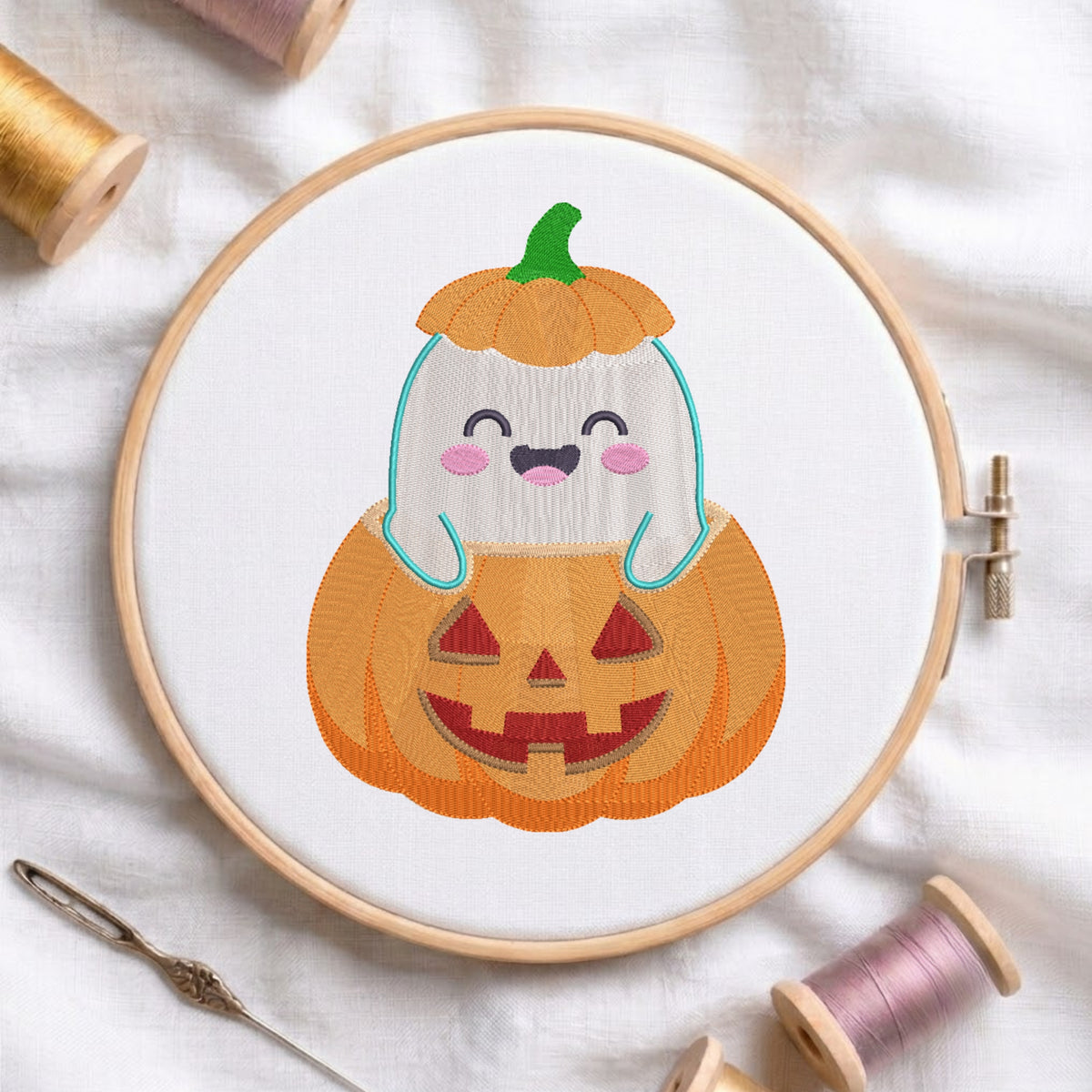 A circular embroidery hoop displays a cute cartoon design of a white ghost with pink cheeks and a green pumpkin stem sitting inside an orange jack-o'-lantern. The hoop is styled on a white fabric background with spools of thread and a needle.
