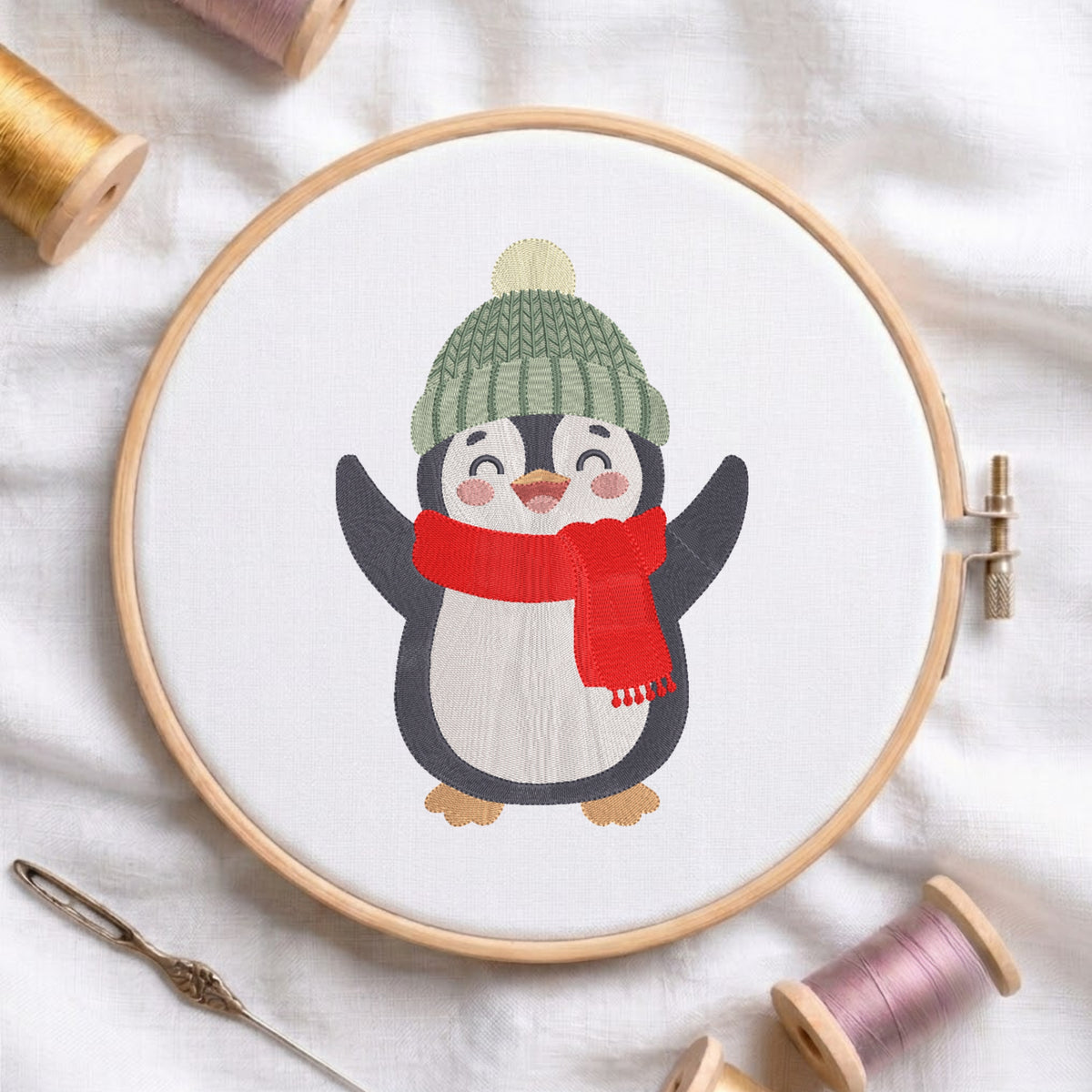A circular embroidery hoop holding white fabric with a stitched penguin design wearing a green knit hat and red scarf, surrounded by spools of thread and a needle on a white fabric background.