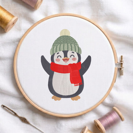 A circular embroidery hoop holding white fabric with a stitched penguin design wearing a green knit hat and red scarf, surrounded by spools of thread and a needle on a white fabric background.