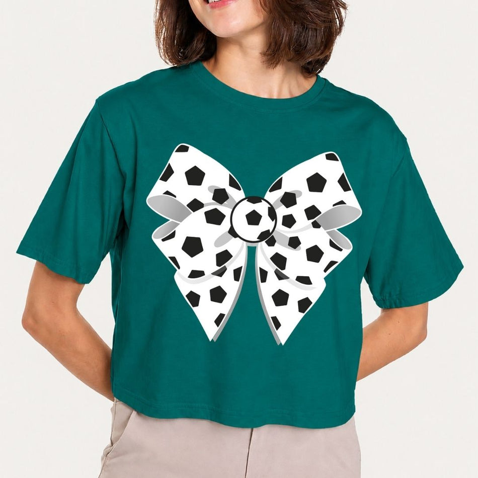 A woman wearing a dark teal short-sleeve crop top with a large white and black soccer-themed bow graphic on the front, paired with light beige pants, against a plain light background.