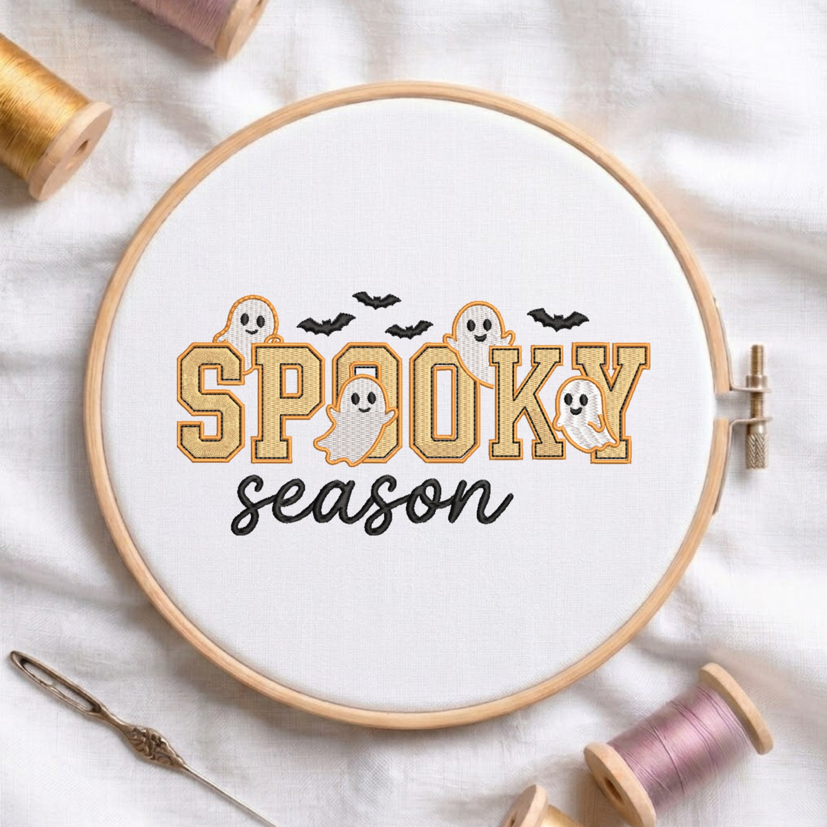 A circular embroidery hoop on white fabric displays the phrase “SPOOKY season” in stitched lettering, with orange outlined letters, black script text, black bats, and small white-and-orange ghost motifs. Spools of thread, a needle, and the hoop are arranged around it on a white fabric background.
