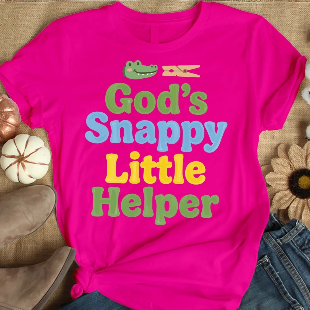 Pink t-shirt with colorful text 'God's Snappy Little Helper' on a textured surface.