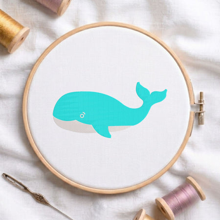 Turquoise blue whale machine embroidery design with a light belly and eye detail, displayed inside a round wooden hoop on white fabric with thread spools and a needle.
